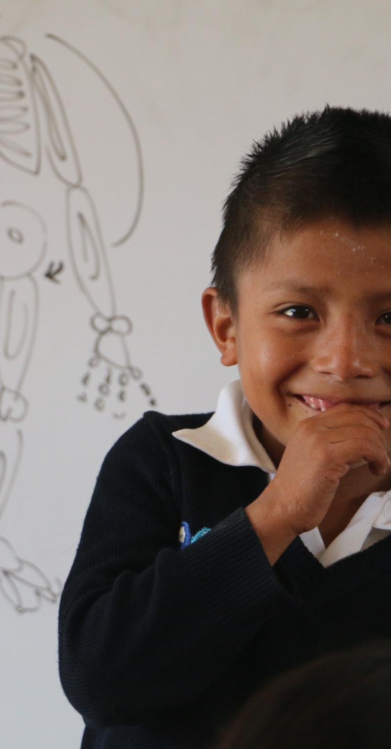A close up of a young boy smiling broadly in front of a white board with a skeleton drawn on it and a word in Spanish that is partially obsured by the boy's head