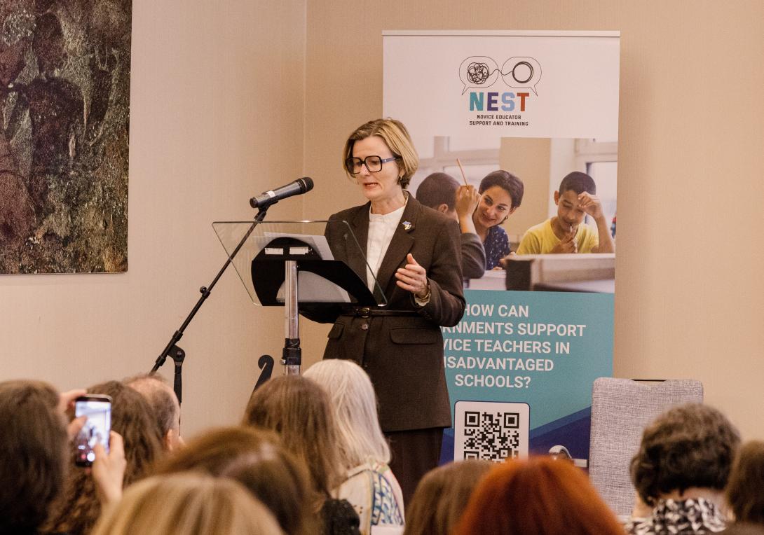 A woman with short blond hair speaks into a microphone at a podium with a pull-poster behind her that says NEST at the top
