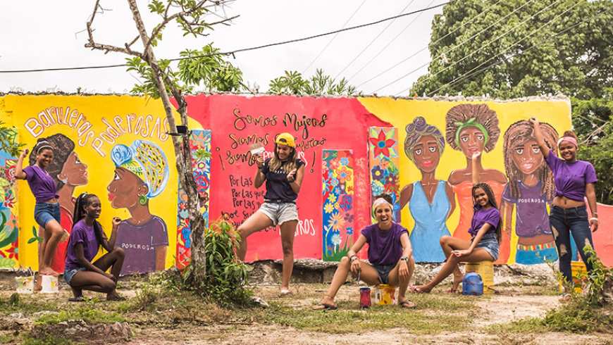 Preview image for the video "Poderosas - Mariana Sanz de Santamaría, Founder Poderosas & Enseña por Colombia Alumna". Preview image for the video "Poderosas - Mariana Sanz de Santamaría, Founder Poderosas & Enseña por Colombia Alumna" - Six young women in purple t-shirts stand in front of a painted mural, which is mostly yellow and red.
