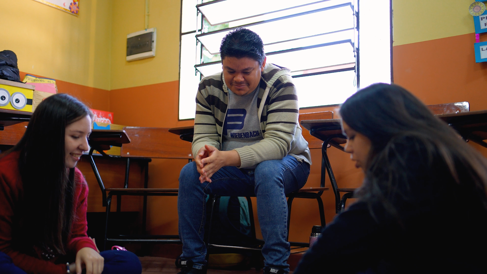 Preview image for the video "Enseña Por Paraguay: Recognizing The Power Within Communities" - A young man with brown hair sits on a chair smiling while looking at something on the floor, where two young women are seated.