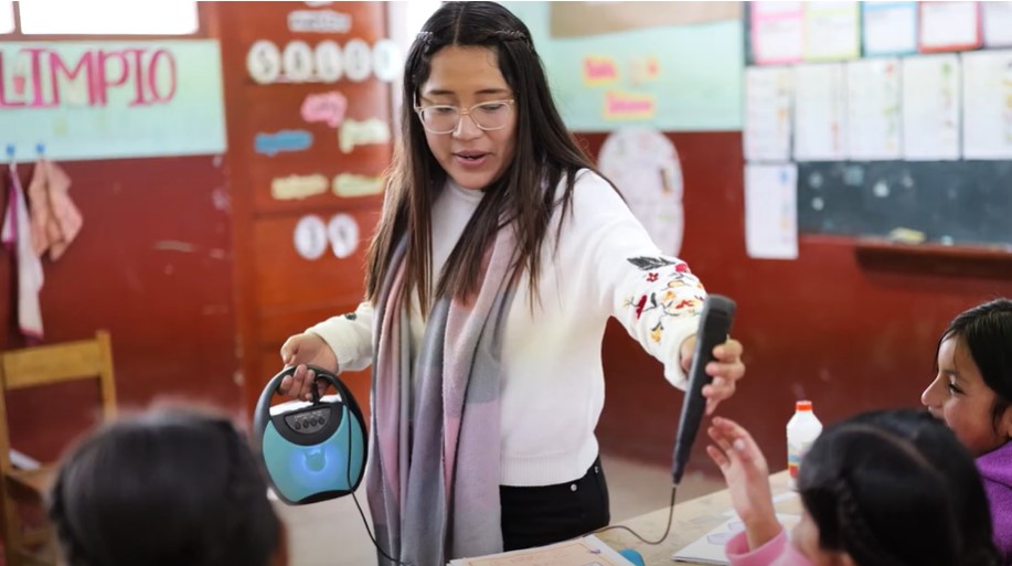 Preview image for the video "Nicole Ames &amp; Students Build a Biodigestor to Tackle Real-World Challenges in Peru" - A young woman with long brown hair stands in a classroom speaking to young children.