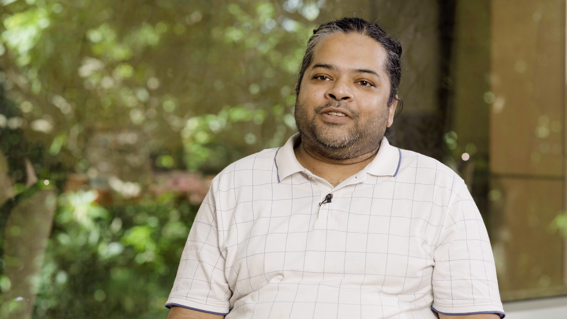 Preview image for the video "Teach For India Alumnus Ashish is Building Positive Childhood Experiences in Conflict Zones" - A medium shot of a man with tan skin and dark, salt-and-pepper hair pulled back, featuring a short beard and mustache. He is wearing a white short-slee polo shirt with a subtle grey grid pattern and a small black lapel microphone clipped to the collar. He is positioned in the center of the frame against a soft, out-of-focus background of lush green foliage and dappled sunlight..