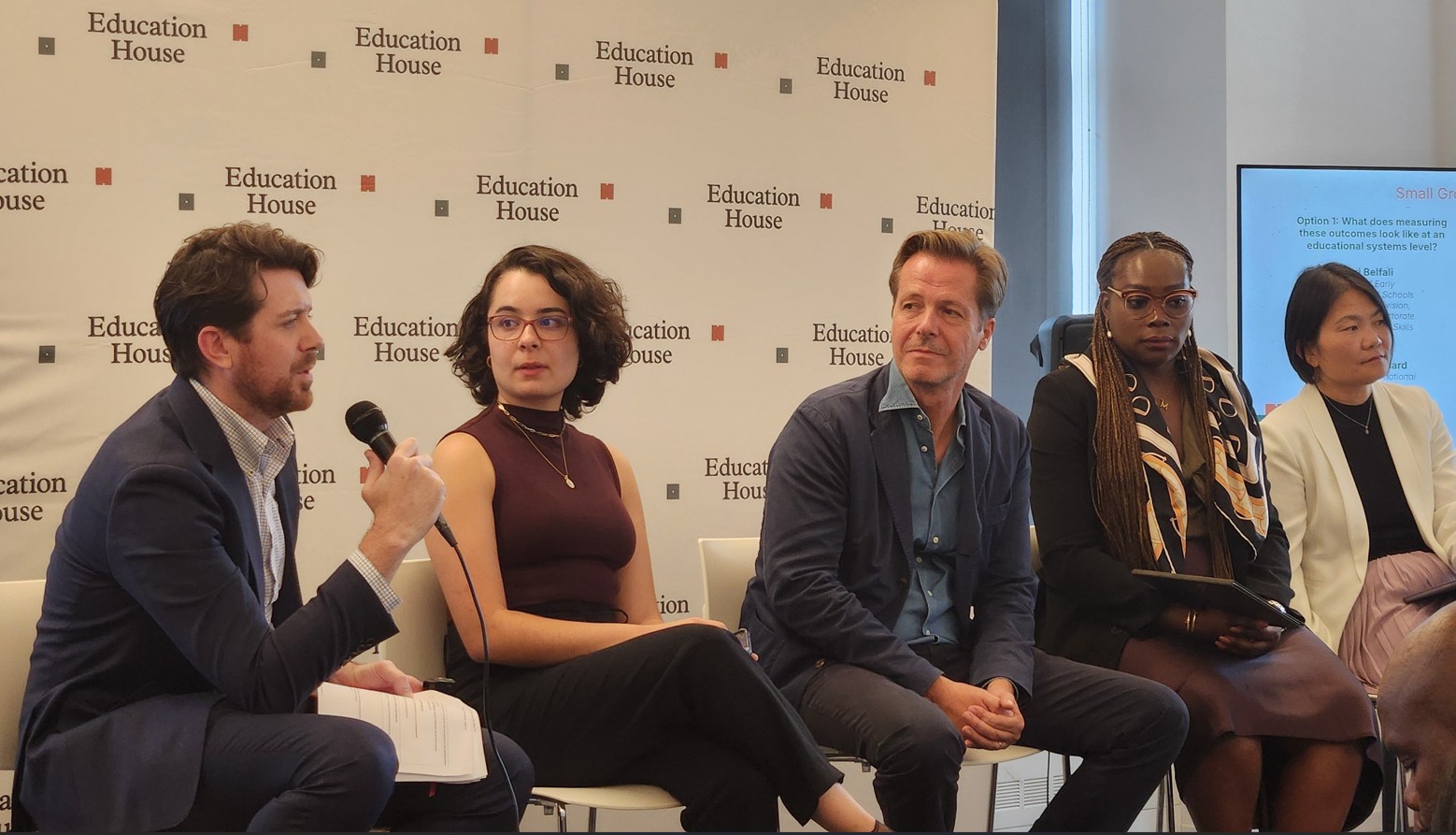 A diverse five-person panel seated on stage in front of an "Education House" backdrop listens as a white man with brown hair and a beard on the far left speaks into a microphone.
