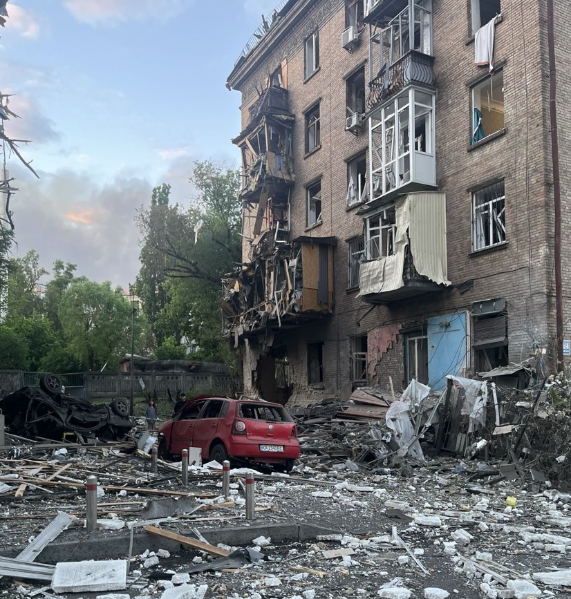 multi-story brick apartment building with extensive blast damage to its exterior and balconies, surrounded by rubble, shattered glass, and damaged cars on the street below.