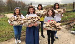 Five smiling girls stand outdoors, each holding a many growing mushrooms