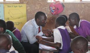 In a clasroom in Uganda, a male teacher wearing a shit and tie speaks with engaged-looking students at their desk