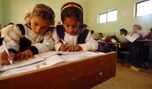 Two young girls write in workbooks at a shared desk in a classroom in Iraq