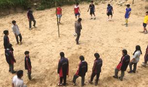 Nepalese children stand outside in a circle around a teacher with a football