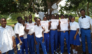 A large group of Nigerian students stand outside with their teacher. They are standing under a tree and holding handwritten signs with messages like "Dream big, work harder" and "Discover your career"