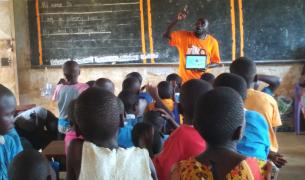 A teacher in an orange Teach For Uganda shirt gesticulates in front of a chalkboard in a classroom full of Ugandan students