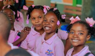 Three Black girls in pink polo shirts with pink bows in their hair smile at the camera