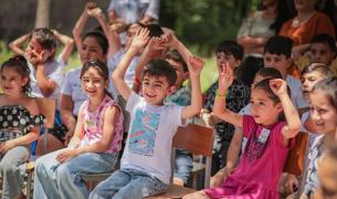 A group of children in colorful casual clothing sitting on chairs smile and some raise both hands above their heads