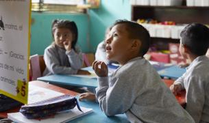 Four young children in school uniforms sit around a desk, the closest to the foreground is a young boy who is looking up at a chart