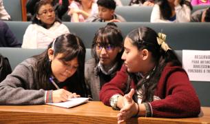 Three Mexican girls in grey and maroon uniforms write on a pad of paper in an auditorium