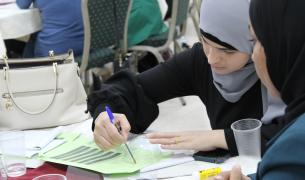 Two women in hijabs sit at a table while one woman traces a circle on a piece of paper