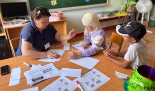 A young female teacher in a blue top shows worksheets to two young students at a table, one wearing a headscarf and the other a baseball cap