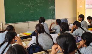 Young girls in school uniforms sit at desks looking at a chalkboard with a web diagram with "ChatGPT" in the middle written on the board