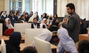 A man with dark hair and brown skin stands amidst a classroom full of girls in headscarfs sitting in front of computer screens and looking at him with attention