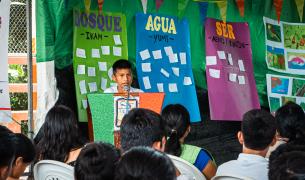 A young boy with tan skin and brown hair speaks at a podium in front of a seated crowd with colorful posters saying "Bosque" "Agua" and "Ser" behind him