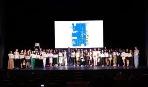 A large group of people on a stage hold up signs, with a large white sign with blue writing behind them