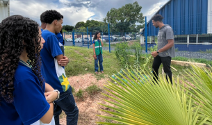 A group of students in blue uniforms stands in a grassy outdoor area, listening to a young man explain a gardening project involving small plants and plastic bottles