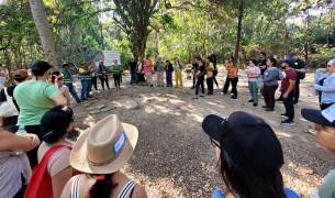 A large, diverse group of people stands in a wide circle in a sun-dappled forest clearing. At the edge of the circle, several individuals in green vests lead a discussion in front of an informational sign. The scene is captured from a wide-angle perspective within the group, surrounded by lush green trees and foliage.
