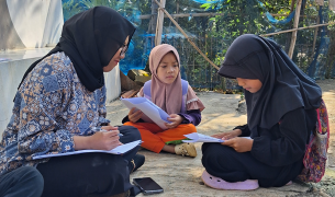An educator wearing a black hijab and a patterned shirt sits on the ground outdoors, reviewing papers with two young schoolgirls who are also wearing hijabs.