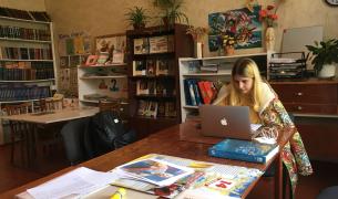 A young woman withe blonde hair and wrapped in a colorful blanket sits at a wooden table working on a laptop inside a classroom library filled with bookshelves.