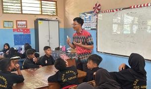 A teacher standing in front of a whiteboard, gesturing while presenting a lesson to students seated around tables in a classroom