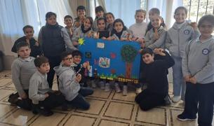A classroom group of school children in grey uniforms posing indoors around a large blue poster project showcasing nature themes, including a tree crafted with bottle caps, butterflies, flowers, and the Earth.