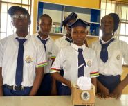 Four Nigerian teens in school uniforms stand behind a camera made out of a cardboard box and other materials