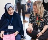 A young student in a hijab sits on the floor next to a blonde, white woman. They are smiling and talking