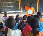 A teacher in an orange Teach For Uganda shirt gesticulates in front of a chalkboard in a classroom full of Ugandan students