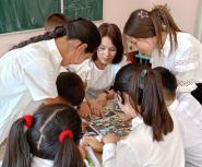 A young female teacher and several teen girls in white shirts, many with their hair in ponytails, look intently at a worksheet on a desk 