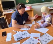 A young female teacher in a blue top shows worksheets to two young students at a table, one wearing a headscarf and the other a baseball cap