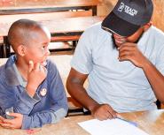 A young Black man in a baseball cap that says Teach For Niger and a young Black boy smile at each other while sharing a wooden desk in a classroom