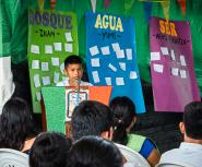 A young boy with tan skin and brown hair speaks at a podium in front of a seated crowd with colorful posters saying "Bosque" "Agua" and "Ser" behind him