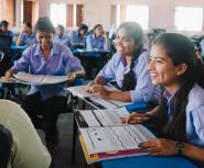 Three you girls sit in desks facing a teacher in a school, they are all smiling