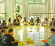 A diverse group of young people and young adults sit in a circle in front of a large window