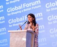 A young woman with long brown hair and tan skin stands at a podium in front of a background that says Global Forum for Shaping a Better Future