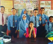 An Asian woman in a pink shirt smiles while seated at table surrounded by teenage students in blue uniforms