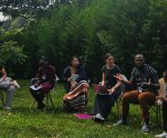 A group of six adults sit in a row on chairs outside on a green grass field in front of some trees