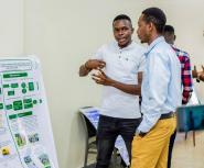An African man stands in front of an academic poster explaining it to another young man while serveral other people surround them