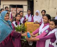 A group of South Asian girls in purple school uniforms with white sashes surrounds a project