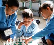 Two young boys and a young girl with brown hair and tan skin in blue school uniforms play chess on the floor