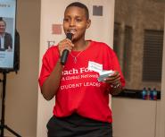 A young african student speaks into a handheld microphone, she is wearing a red shirt that says "Teach For All student leader"