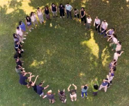 A light skinned young man with brown hair and a beard demostrates a science experiment using beekers to a diverse group of teenagers