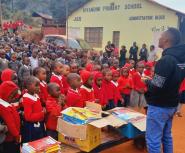 A young Black man stands in front of a bench piled with workbooks and a large crowd of Black children in school uniforms outside of a school
