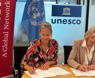 Two white women with greying blonde hair sit side by side signing papers in front of them, a red Teach For All banner on the right, and a UNESCO logo above one's head