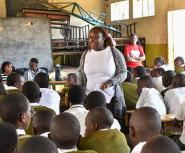 A young African woman in a white shirt and grey sweater stands in the middle of a classroom full of students